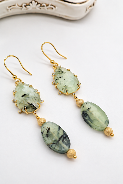 Dettaglio di orecchini pendenti in prehnite naturale con cabochon sfaccettato, zirconi e pietre ovali lisce, finiture dorate.