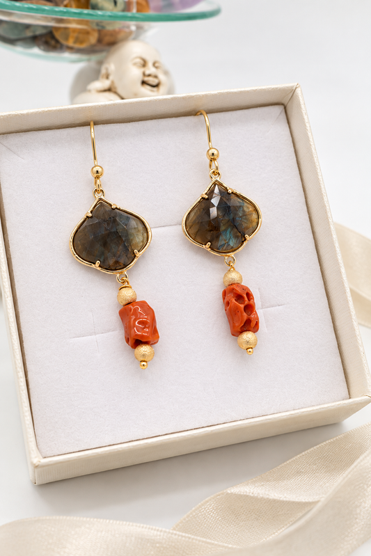 Orecchini pendenti in labradorite grigia sfaccettata con riflessi blu iridescenti, montatura dorata e corallo mediterraneo arancione, fotografati su sfondo chiaro.